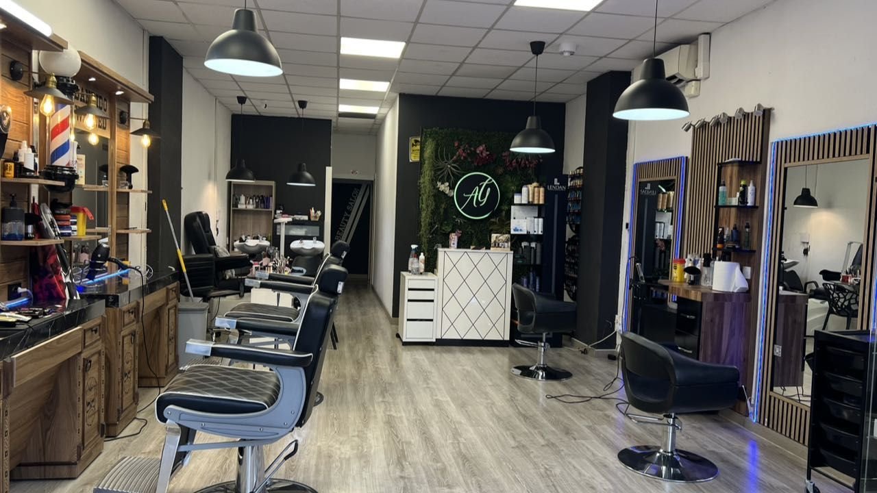 Sala principal de AG Beauty Salon: zona de barbería a la izquierda, lavacabezas y tocadores de peluquería a la derecha, con el logo AG en neón sobre pared vegetal